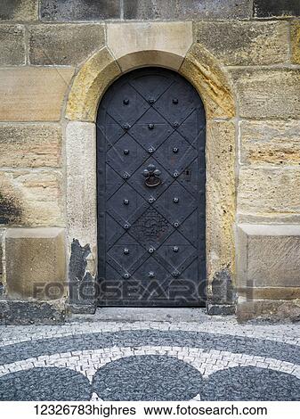 Stock Photograph - An arched metal door in a stone block wall with decorative paving stones in a design; Prague, Czech Republic. Fotosearch