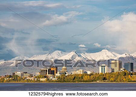 Stock Photograph - Downtown Anchorage as seen from Point Woronzof, atmospheric clouds shrouding the Chugach Mountains in the background, autumn coloured trees on the shoreline, South-central Alaska; Anchorage, Alaska, United States of America. Fotosearch
