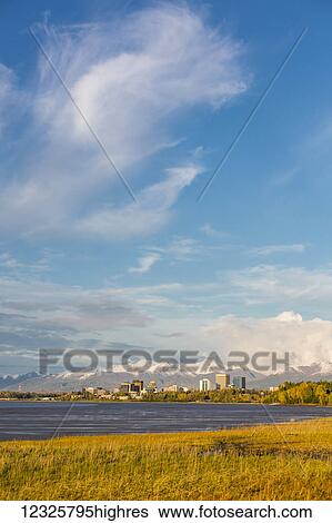 Stock Photograph - Downtown Anchorage as seen from Point Woronzof, atmospheric clouds shrouding the Chugach Mountains in the background, autumn coloured grasses line the shoreline in the foreground, South-central Alaska; Anchorage, Alaska, United States of America. Fotosearch