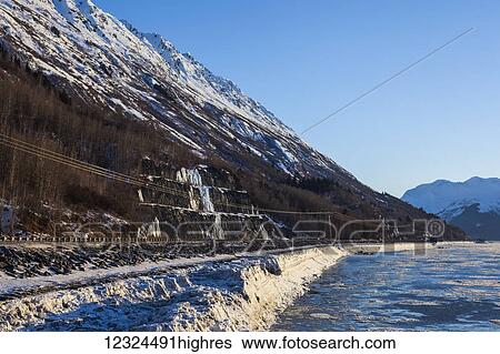 Stock Photograph - Early morning winter view along Turnagain Arm showing the rugged Chugach Mountains cutting down into the ice covered water while Seward Highway and the railroad cut a thin line between them; Alaska, United States of America. Fotosearch