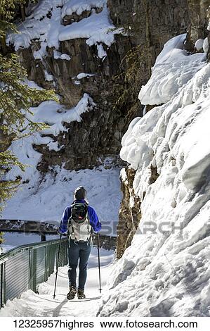 Stock Photograph - Female hiker on snow covered trail along snow covered rock cliff and fencing, Johnson Canyon; Alberta, Canada. Fotosearch