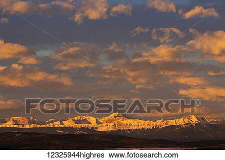 Glowing warmly lit snow covered mountain range and foothills with glowing clouds and blue sky at sunrise; Calgary, Alberta, Canada View Large Photo Image Stock Photograph - Glowing warmly lit snow covered mountain range and foothills with glowing clouds and blue sky at sunrise; Calgary, Alberta, Canada. Fotosearch