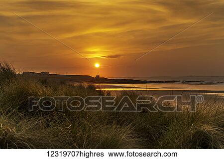 Stock Photograph - Golden sun setting in an orange sky along the coast with Bamburgh Castle in the distance; Bamburgh, Northumberland, England. Fotosearch