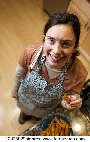 High angle view of a woman in a kitchen wearing an apron and holding a piece of baked sweet potato; Alaska, United States of America View Large Photo Image Stock Photograph - High angle view of a woman in a kitchen wearing an apron and holding a piece of baked sweet potato; Alaska, United States of America. Fotosearch