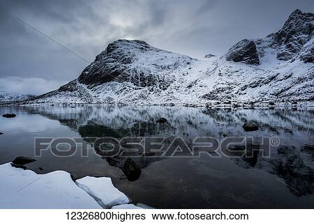 Stock Photograph - Landscape of rugged, snow covered mountains reflected in tranquil ocean water; Lofoton Islands, Nordland, Norway. Fotosearch