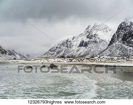 Stock Photograph - Landscape of the coastline and rugged, snow covered mountains under a cloudy sky; Lofoten Islands, Nordland, Norway. Fotosearch