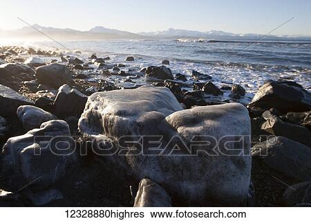 Stock Photograph - Large rocks along the shore with a view of mountains along the coastline; Alaska, United States of America. Fotosearch