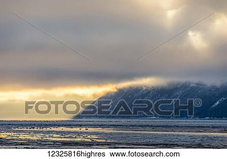 Stock Photograph - Low altitude clouds are cast in warm sunset light along Turnagain Arm and the Seward Highway in winter, sea ice covering the ocean in the foreground, the Chugach Mountains revealed in the background, South-central Alaska; Alaska, United States of America. Fotosearch