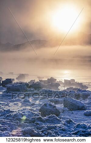 Stock Photograph - Low altitude fog is cast in warm sunset light along Turnagain Arm and the Seward Highway in winter, sea ice covering the ocean in the foreground, the Chugach Moutains revealed in the background, South-central Alaska; Alaska, United States of America. Fotosearch