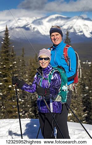 Stock Photograph - Male and female cross country skiiers on a lookout with snow covered mountains, blue sky and clouds in the background; Lake Louise, Alberta, Canada. Fotosearch