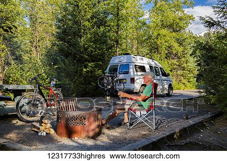 Stock Photograph - Male camper enjoying a fire in Williwaw USFS Campground with a bicycle and Sportsmobile camper van in the background in Portage Valley, Summer, Alaska, USA. Fotosearch