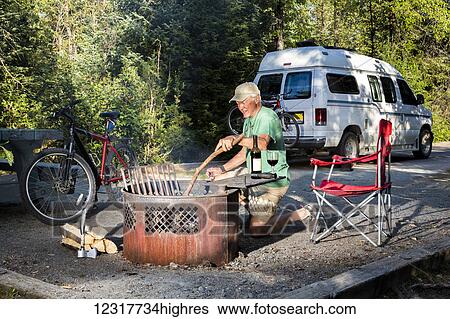 Stock Photograph - Male camper enjoying a fire in Williwaw USFS Campground with a bicycle and Sportsmobile camper van in the background in Portage Valley, Summer, Alaska, USA. Fotosearch