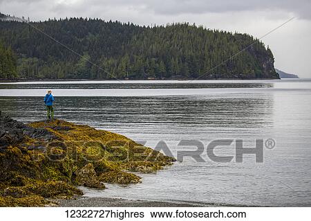 Stock Photograph - Man standing on a rock overlooking the ocean, Gwaii Haanas National Park; British Columbia, Canada. Fotosearch