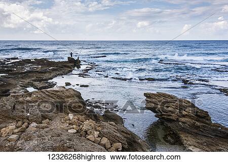 Stock Photograph - Men stand fishing on rock at the water's edge; Caesarea, Haifa District, Israel. Fotosearch