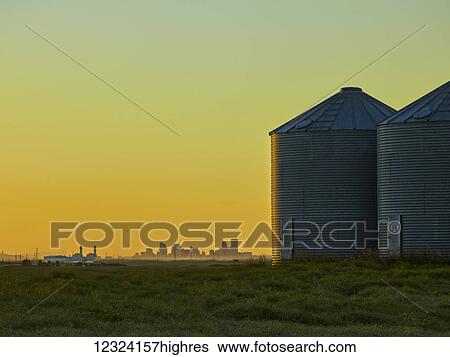 Stock Photograph - Metal silos on a farm field with the skyline of a city in the distance at sunset, near Calgary; Alberta, Canada. Fotosearch