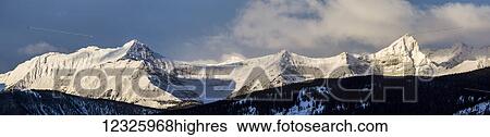 Stock Photograph - Panorama of snow covered mountains with early morning light, silhouetted forest in the foreground, blue sky and clouds; Kananaskis Country, Alberta, Canada. Fotosearch