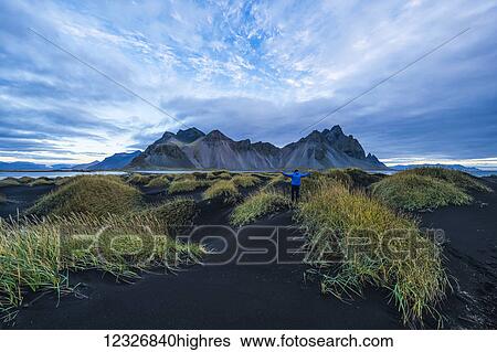 Stock Photograph - Person standing on a black sand mound looking at Vestrahorn or Stokknes on the South coast of Iceland; Hofn, Iceland. Fotosearch