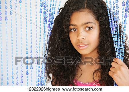 Stock Photograph - Portrait of a young girl with dark, curly hair and brown eyes standing and holding strings of a beaded curtain; Vancouver, Washington, United States of America. Fotosearch