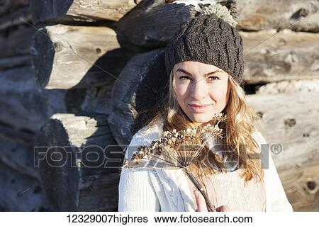 Stock Photograph - Portrait of a young woman holding a sprig of dried wildflowers by a log cabin; Homer, Alaska, United States of America. Fotosearch