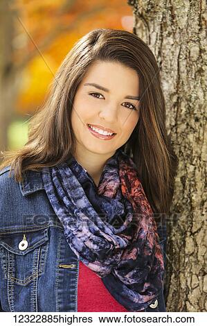 Stock Photograph - Portrait of a young woman standing against a tree trunk with autumn colours in the background; Oregon, United States of America. Fotosearch