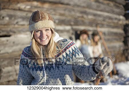Stock Photograph - Portrait of a young woman wearing a hat and sweater by a log cabin in winter; Homer, Alaska, United States of America. Fotosearch