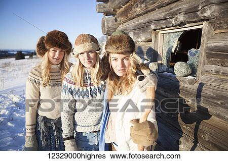 Stock Photograph - Portrait of three girls standing by a log cabin wearing fur hats; Homer, Alaska, United States of America. Fotosearch