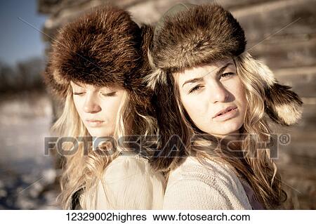 Stock Photograph - Portrait of two girls standing by a log cabin wearing fur hats; Homer, Alaska, United States of America. Fotosearch