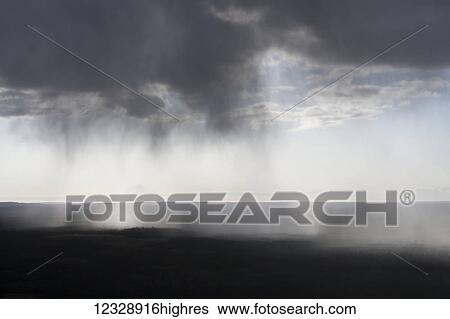 Stock Photograph - Rain falls from dark heavy clouds to the dark land below with the horizon through the mist of rain; Alaska, United States of America. Fotosearch