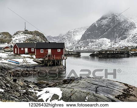 Stock Photograph - Red buildings along the water's edge under a cloudy sky with snow on the mountains; Lofoten Islands, Nordland, Norway. Fotosearch