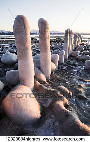 Stock Photograph - Rocks and pilings covered with snow and ice along the shore at the water's edge, with a view of the coastline and mountains in the distance; Alaska, United States of America. Fotosearch
