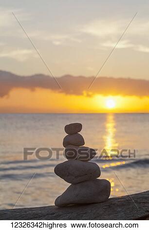Stock Photograph - Rocks balanced in a pile on the shore as the golden sun sets over the water and horizon; Honolulu, Oahu, Hawaii, United States of America. Fotosearch