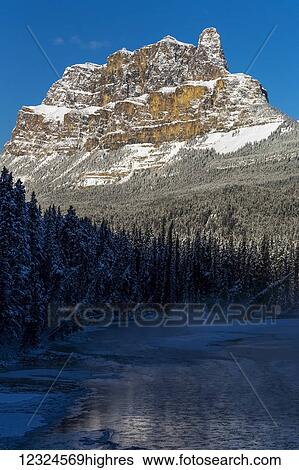 Rugged snow covered mountain reflecting off open water on a river with frosted and snow covered evergreen trees with blue sky; Banff, Alberta, Canada View Large Photo Image Stock Photograph - Rugged snow covered mountain reflecting off open water on a river with frosted and snow covered evergreen trees with blue sky; Banff, Alberta, Canada. Fotosearch