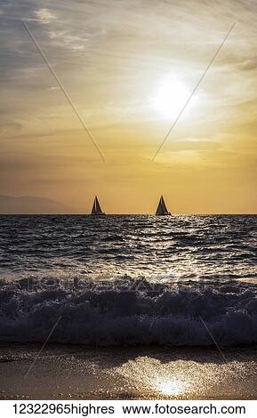 Stock Photograph - Sailboats sail as the sun sets in the Bay of Banderas; Puerto Vallarta, Mexico. Fotosearch