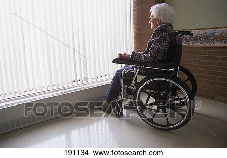 Picture - Senior woman on wheelchair looking out of window with blinds. Fotosearch