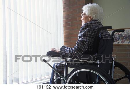 Stock Image - Senior woman on wheelchair looking out of window with blinds. Fotosearch