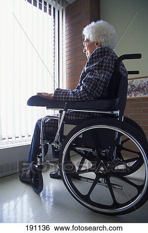 Stock Photograph - Senior woman on wheelchair looking out of window with blinds. Fotosearch