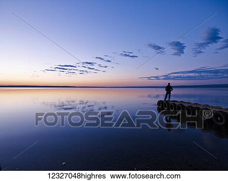 Silhouette of a person standing at the end of a dock looking out over a tranquil lake at sunset, Gregoire Lake Provincial Park; Alberta, Canada View Large Photo Image Stock Photograph - Silhouette of a person standing at the end of a dock looking out over a tranquil lake at sunset, Gregoire Lake Provincial Park; Alberta, Canada. Fotosearch