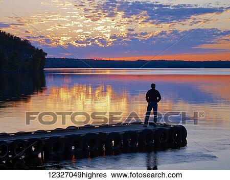 Silhouette of a person standing at the end of a dock looking out over a tranquil lake at sunset, Gregoire Lake Provincial Park; Alberta, Canada View Large Photo Image Stock Photograph - Silhouette of a person standing at the end of a dock looking out over a tranquil lake at sunset, Gregoire Lake Provincial Park; Alberta, Canada. Fotosearch