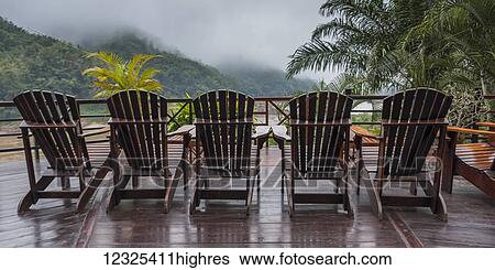 Stock Photograph - Six wooden adirondack chairs sit in a row on a wooden deck with a view of the low cloud over a mountainous landscape; Oudomxay Province, Laos. Fotosearch