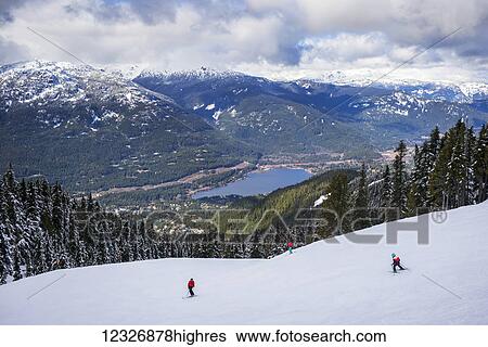 Stock Photograph - Skiers and snowboarders making their way down a ski hill with a magnificent view of a lake and the Canadian Rocky Mountains; Whistler, British Columbia, Canada. Fotosearch