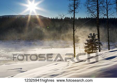 Stock Photograph - Snow covered hillside along a river with fog and steam rising and a sunburst with blue sky, West of Bragg Creek; Alberta, Canada. Fotosearch