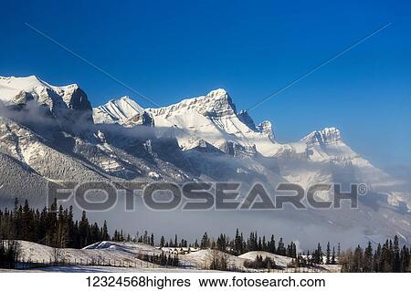 Stock Photograph - Snow covered mountain range with clouds in the valley, frosted trees and blue sky; Canmore, Alberta, Canada. Fotosearch