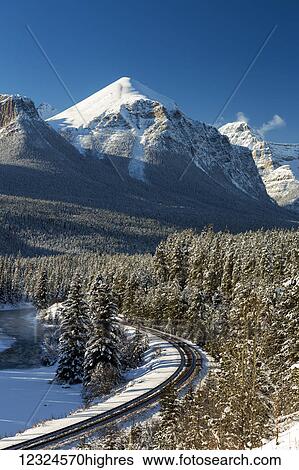 Stock Photograph - Snow covered mountain range with frosted and snow covered evergreen trees along a river with train tracks and blue sky; Banff, Alberta, Canada. Fotosearch
