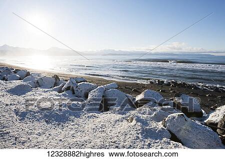 Stock Photograph - Snow covered rocks and beach with a view of the ocean and mountains along the coastline; Alaska, United States of America. Fotosearch