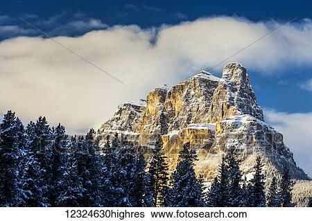 Stock Photograph - Snow covered rugged mountain and evergreen trees with blue sky and clouds; Banff, Alberta, Canada. Fotosearch