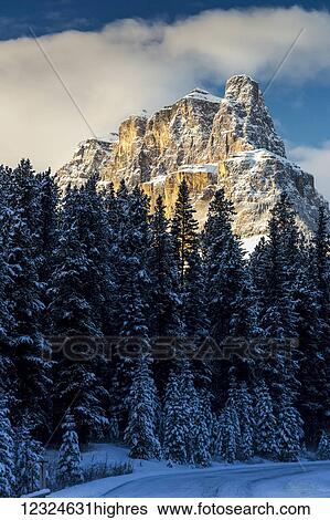 Stock Photograph - Snow covered rugged mountain and evergreen trees, snow covered road, with blue sky and clouds; Banff, Alberta, Canada. Fotosearch