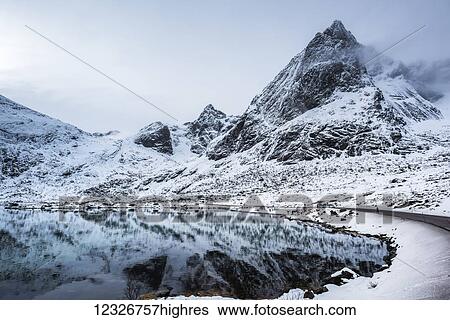 Stock Photograph - Snowy landscape of rugged mountains reflected in tranquil water; Lofoten Islands, Nordland, Norway. Fotosearch