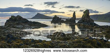 Stock Photograph - Sunrise over some lava pillars with a large sea cliff rising in the horizon, Strandir Coast; West Fjords, Iceland. Fotosearch