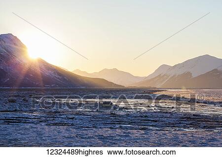 Stock Photograph - Sunrise peaks over the Chugach Mountains into Turnagain Arm on an icy winter morning; Alaska, United States of America. Fotosearch
