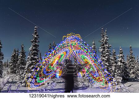 Stock Photograph - The Aurora Borealis dances over the top of a vibrant spiral light painting, the blurry figure of a man in the middle of the light painting, moonlight casting shadows on s0w covered spruce trees on a clear winter night, interior Alaska; Gakona, Alaska,.... Fotosearch